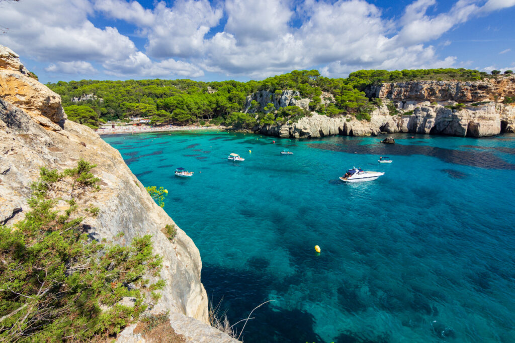 Panoramablick auf eine türkisfarbene Bucht auf Menorca mit Booten, hellen Klippen, Pinienwald und Sandstrand