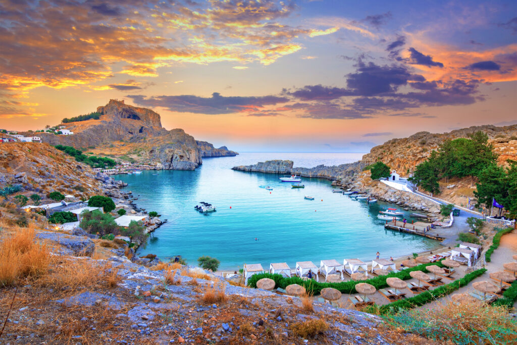 Bucht bei Lindos auf Rhodos mit Booten, Felsen, weißer Kapelle und Sonnenuntergang.