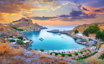 Bucht bei Lindos auf Rhodos mit Booten, Felsen, weißer Kapelle und Sonnenuntergang.