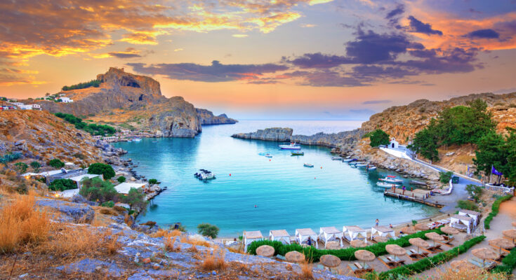 Bucht bei Lindos auf Rhodos mit Booten, Felsen, weißer Kapelle und Sonnenuntergang.