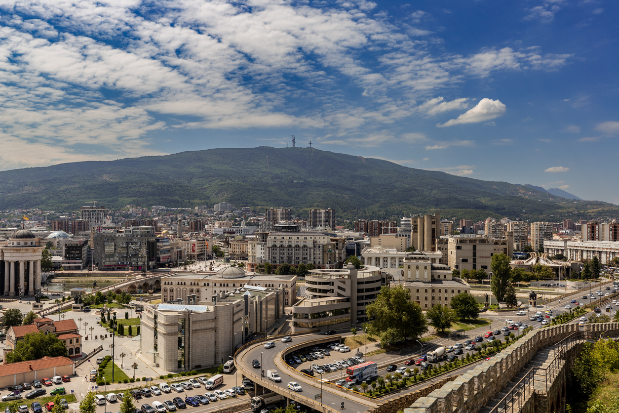 Panorama von Skopje: Blick von der Festung Kale über die Innenstadt mit Brücken über den Vardar, neoklassizistischen Gebäuden und dem bewaldeten Berg Vodno mit Millenniumskreuz im Hintergrund unter leicht bewölktem Himmel.