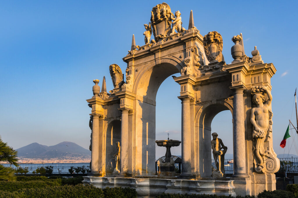Neapel, barocker Brunnen Fontana del Gigante mit drei Bögen und Figuren an der Uferpromenade, Vesuv im Hintergrund, warmes Abendlicht, klarer Himmel
