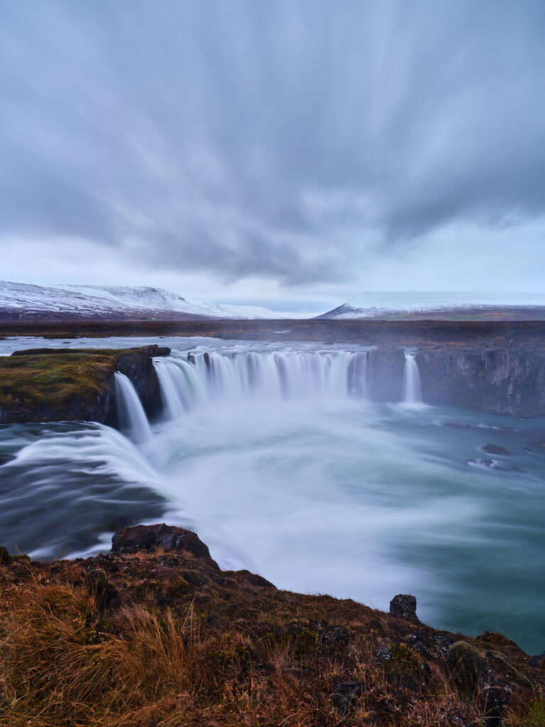 Goðafoss-Wasserfall in Island mit Langzeitaufnahme, schäumendes Wasser, Grasvordergrund und schneebedeckte Hügel.