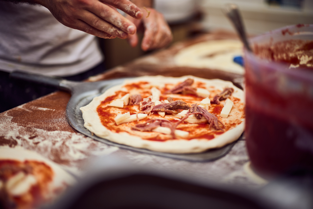 Pizza Napoletana im Entstehen, ausgerollter Teig mit Tomatensauce und Käsewürfeln auf einer Pizzaschaufel, bemehlte Arbeitsfläche, Hände des Pizzaiolo