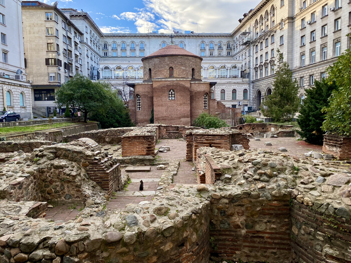 Sofia, Rotunde des Heiligen Georg aus rotem Ziegel, römische Ruinen im Vordergrund, umgeben von mehrgeschossigen Verwaltungsgebäuden, teils bewölkter Himmel