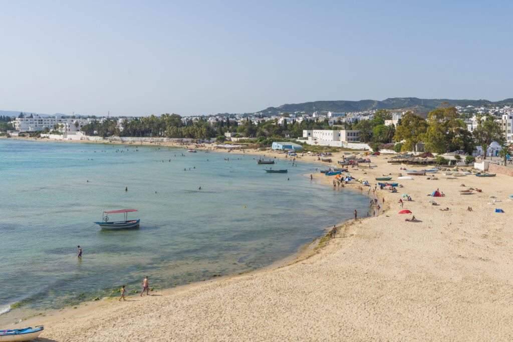 Breiter Sandstrand von Hammamet mit türkisfarbenem, flachem Meer, Badenden, kleinen Booten und Hotels im Hintergrund.