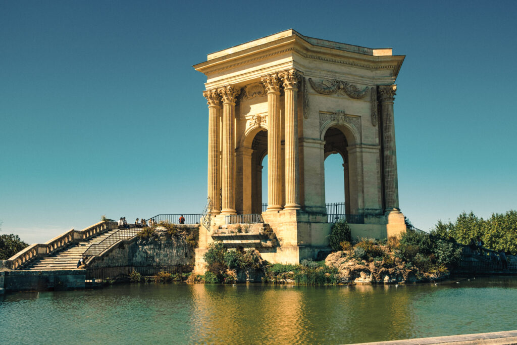 Neoklassischer Pavillon mit korinthischen Säulen auf einer Insel im Wasserbecken der Promenade du Peyrou in Montpellier; links eine breite Freitreppe, einige Besucher und klarer blauer Himmel.