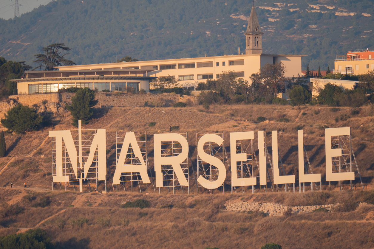 Großer weißer „MARSEILLE“-Schriftzug im Hollywood-Stil auf einem trockenen Hügel, dahinter moderne Gebäude und eine Kapelle vor bewaldeten Hängen.