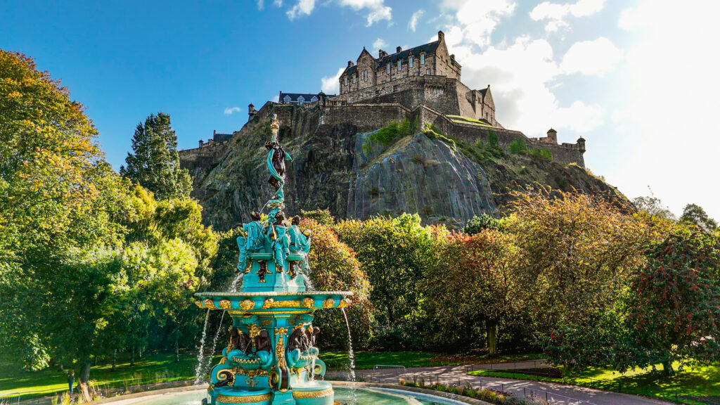 Edinburgh, Castle auf dem Castle Rock mit dem Ross Fountain im Vordergrund, umgeben von Bäumen und Parkwegen, Sonne mit einzelnen Wolken