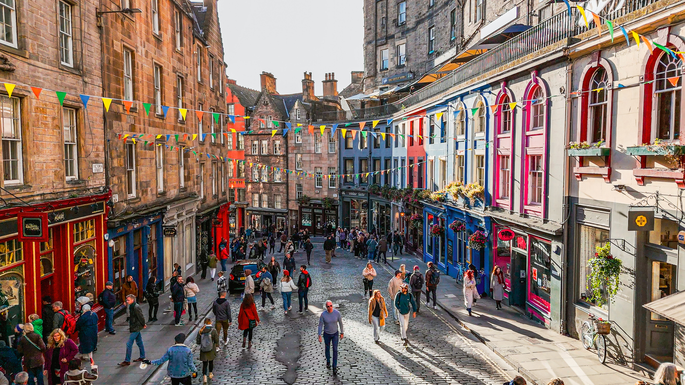 Edinburgh, Victoria Street in der Altstadt mit farbigen Ladenfassaden, Wimpelketten über der Straße, Kopfsteinpflaster und vielen Fußgängern bei Tageslicht