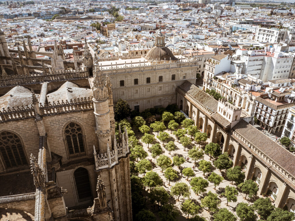 Sevilla, Patio de los Naranjos neben der Kathedrale, regelmäßige Reihen von Orangenbäumen mit umlaufenden Arkaden, vorn gotische Strebwerke und Fialen, Blick von oben über die Altstadt