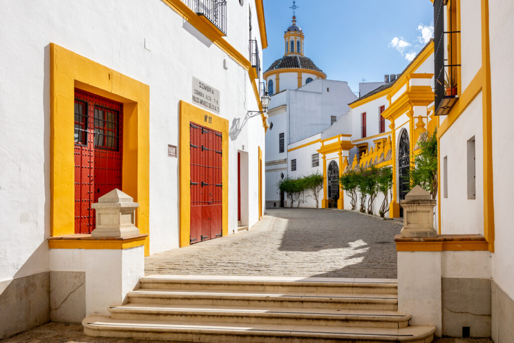 Sevilla, gepflasterte Gasse mit weiß verputzten Fassaden und gelben Laibungen, rote Holztore und schmiedeeiserne Balkone, im Hintergrund die Kuppel eines Barockbaus, sonniger Tag