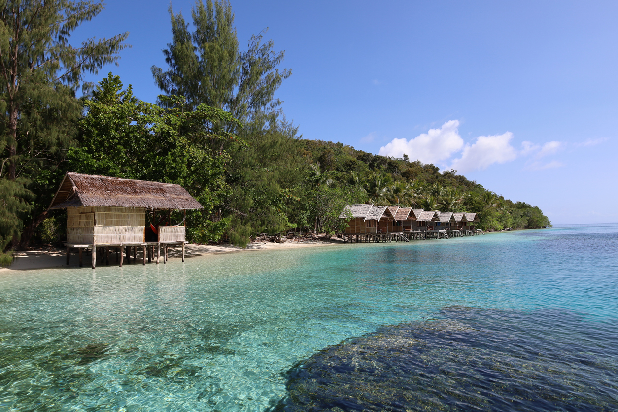 Stelzenhütten über klarem, türkisfarbenem Wasser vor bewaldetem Ufer in Raja Ampat.