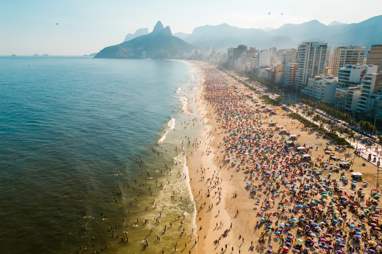 Luftaufnahme: voller Ipanema-Strand mit bunten Sonnenschirmen; Meer links, Morro Dois Irmãos im Dunst.