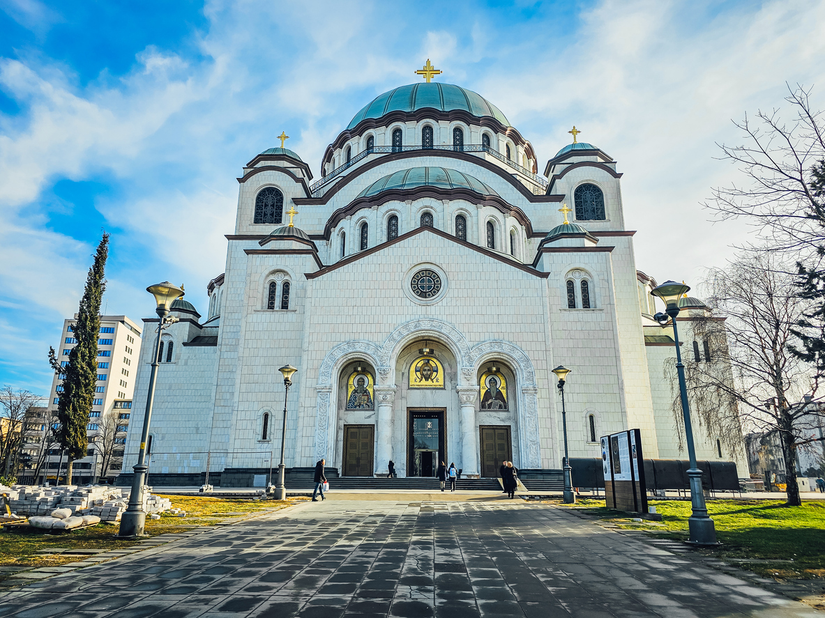 Belgrad, Kirche des Heiligen Sava frontal, weiße Fassade mit drei Portalen und Mosaiken, kupfergrüne Kuppeln mit goldenen Kreuzen, Vorplatz mit Laternen und Passanten, blauer Himmel