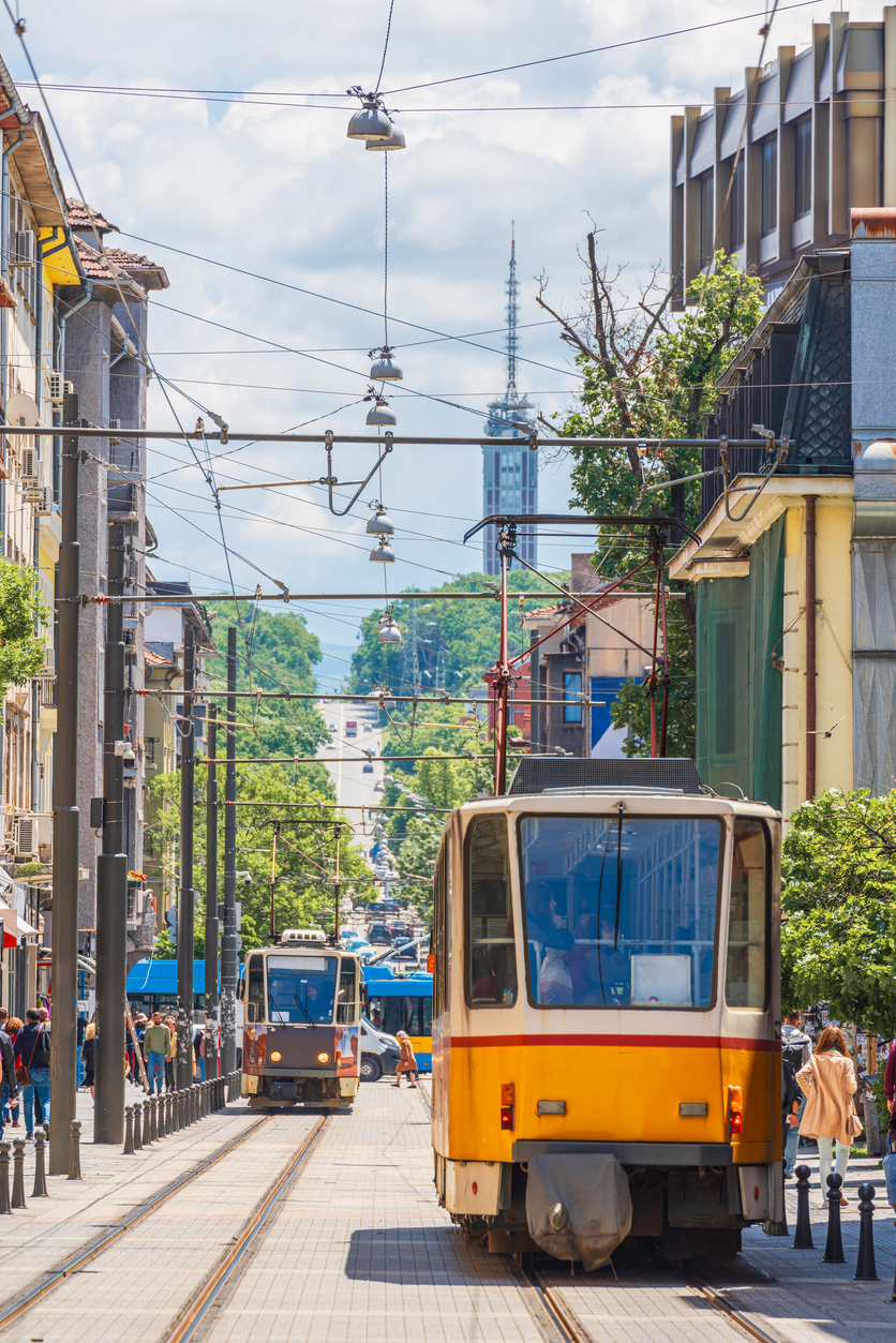 Sofia, gelbe Straßenbahn im Vordergrund auf Innenstadtgleisen, zweite Tram entgegenkommend, dichte Oberleitungen, Fußgänger an den Seiten, Antennenturm und Hügellandschaft im Hintergrund, sonniger Tag