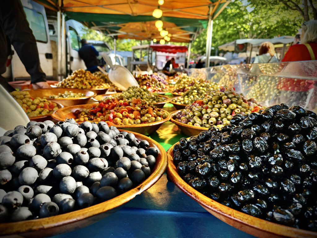 Nahaufnahme von Schalen mit schwarzen und grünen Oliven auf einem Wochenendmarkt im 2. Arrondissement von Lyon; Marktstände unter farbigen Planen am Saône-Ufer, Besucher im Hintergrund.
