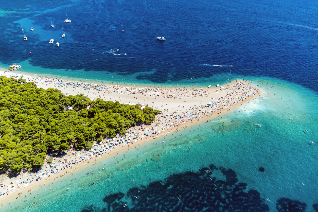 Luftaufnahme von Zlatni Rat, pfeilförmige Kieselspitze mit Kiefernwald, vielen Strandbesuchern und Booten im türkisblauen Meer