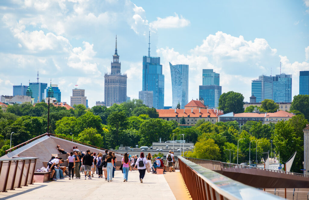 Menschen auf erhöhter Promenade; Warschauer Skyline mit Kulturpalast und modernen Hochhäusern, viel Grün.