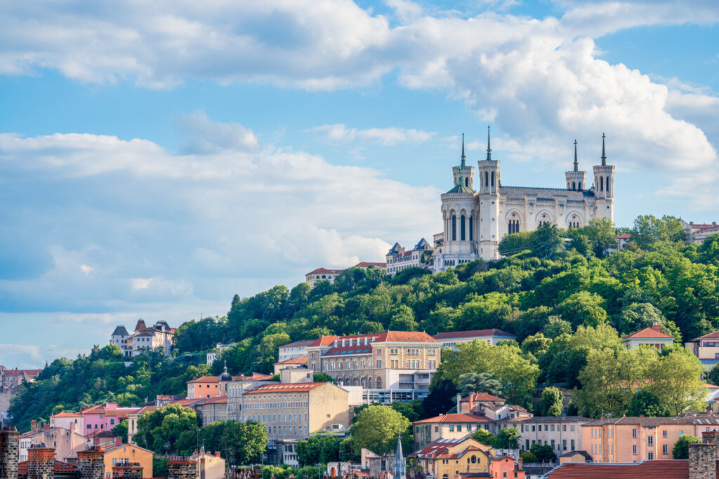Panoramablick auf die Basilika Notre-Dame de Fourvière, die auf dem grünen Fourvière-Hügel über den historischen Häusern Lyons thront, unter leicht bewölktem Himmel.