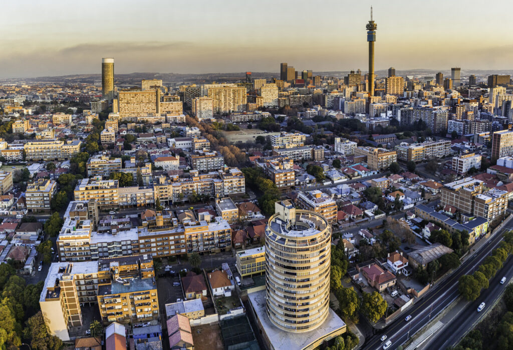 Luftaufnahme von Johannesburg mit Hillbrow-Turm, dicht bebauten Vierteln und zylindrischem Hochhaus im Abendlicht.