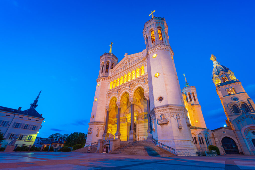 Weitwinkelaufnahme der Basilika Notre-Dame de Fourvière in Lyon bei Abenddämmerung; goldgelb beleuchtete romanisch-byzantinische Fassade mit vier Türmen, breite Treppen und klarer Himmel.