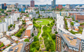 Luftaufnahme des Park-Central-Viertels in Birmingham mit kreisförmiger Rasenfläche, geschwungenen Fußwegen und modernen Apartmenthäusern; dahinter die Skyline unter bewölktem Himmel.