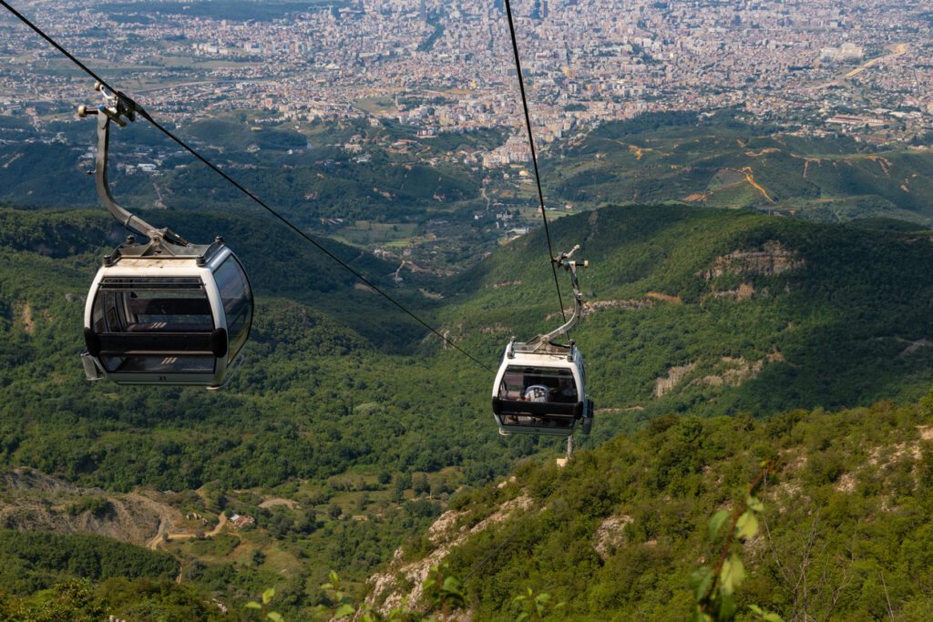 Zwei Seilbahngondeln schweben über den grünen Hängen des Berges Dajti, im Hintergrund breitet sich die Stadt Tirana aus, klare Sicht an einem sonnigen Tag