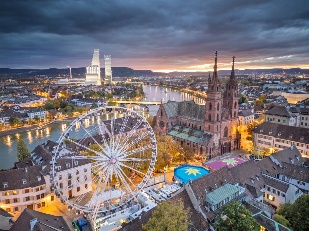 Luftaufnahme von Basel bei Dämmerung mit Basler Münster, Riesenrad am Münsterplatz und dem Rhein mit Brücken und Stadtlichtern