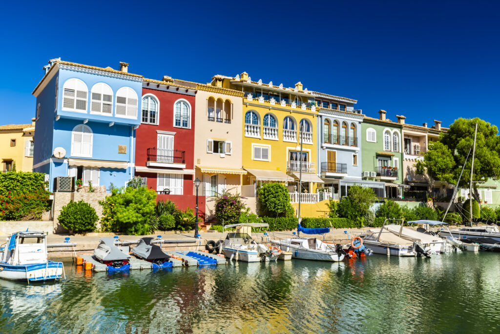 Reihe farbenfroher mediterraner Häuser mit Booten am ruhigen Kanal der Marina Port Saplaya bei Valencia unter tiefblauem Himmel.