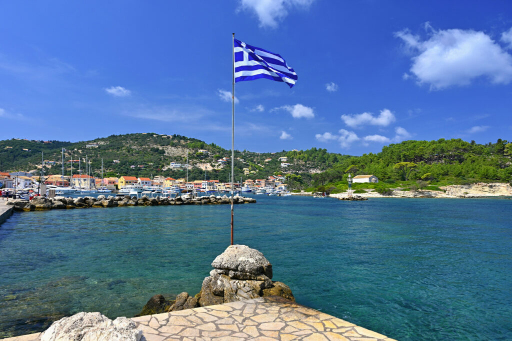 Griechische Flagge am Pier im Hafen von Paxos, klare Bucht, Yachten, bunte Häuser und grüne Hügel.
