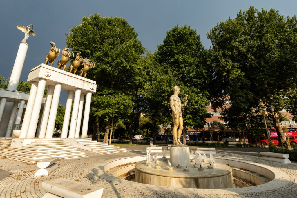 Runder Brunnen mit männlicher Statue in Skopje, daneben eine weiße Kolonnade mit goldener Quadriga; hoher Baumbestand und dramatisch dunkler Himmel im Hintergrund.