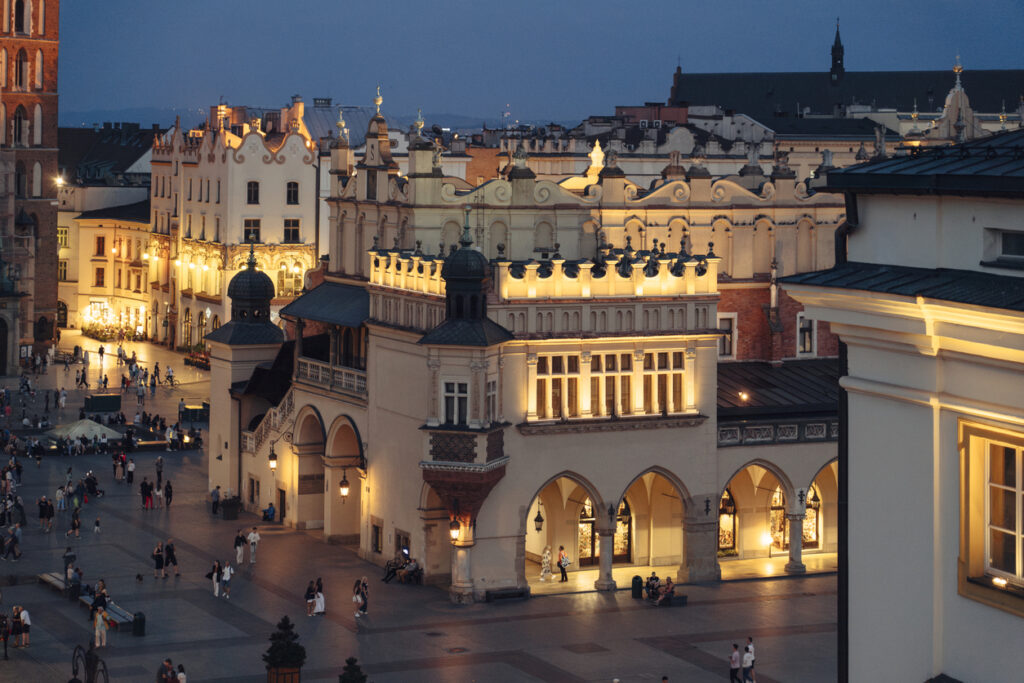 Beleuchtete Tuchhallen (Sukiennice) auf dem Hauptmarkt in Krakau bei Nacht, mit Menschen, die über den Platz schlendern.