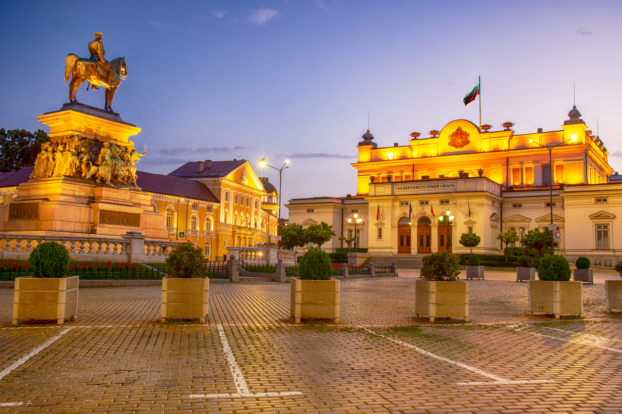 Sofia, Parlamentsgebäude der Nationalversammlung in warmem Abendlicht, links Reiterdenkmal, gepflasterter Platz mit Kübeln und Bepflanzung, klarer Himmel