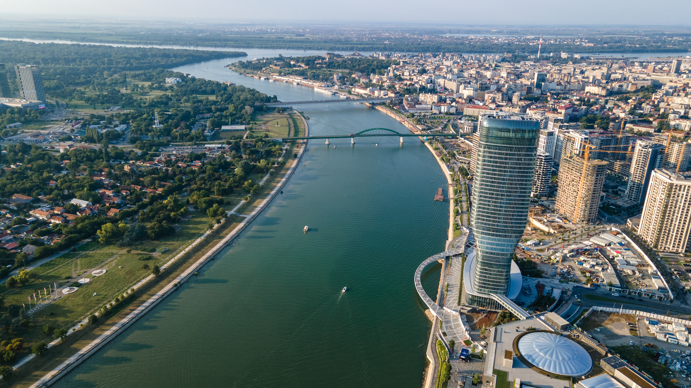 Belgrad, Luftaufnahme des Sava-Ufers mit dem geschwungenen Hochhaus Kula Belgrade am Belgrade Waterfront, grüner Bogen der Alten Sava-Brücke, Promenade und Uferparks, Boote auf dem Fluss, klarer Himmel