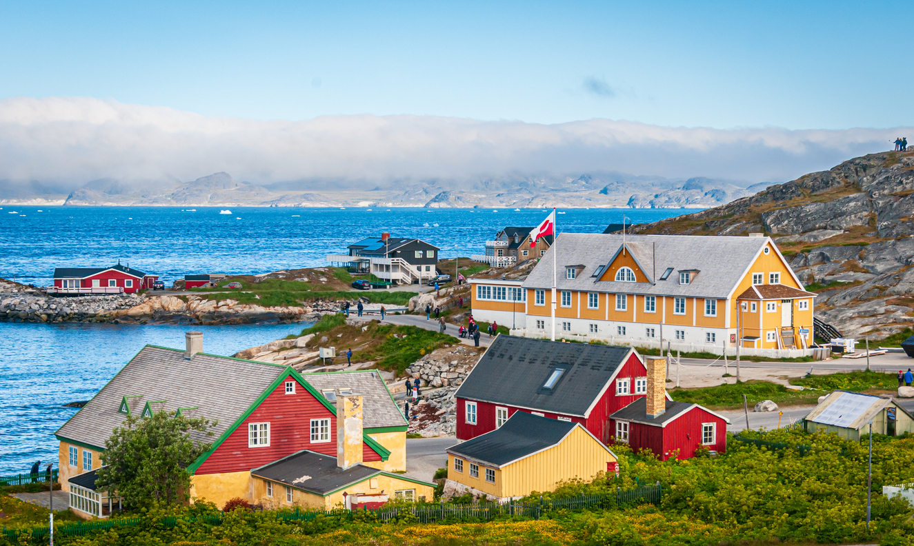 Bunte Holzhäuser am Fjord; großes gelbes Gebäude mit Flagge; Meer, Inseln und Nebelbank im Hintergrund.