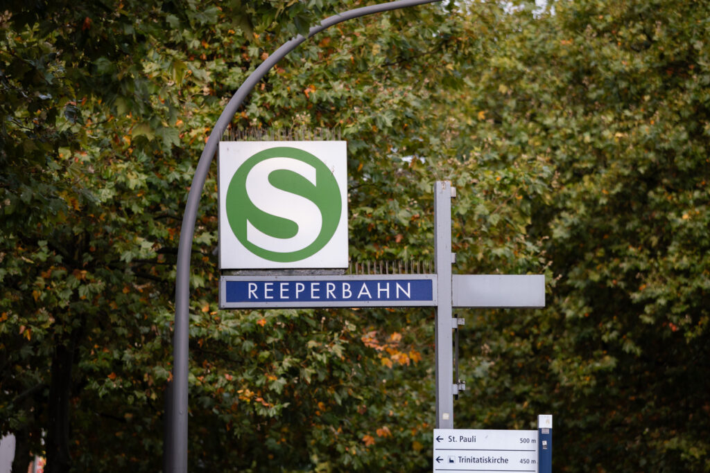 Hamburg, S-Bahn-Logo und Stationsname „Reeperbahn“ an einem Mastschild, darunter Wegweiser zu St. Pauli und Trinitatiskirche, dichter Laubbestand im Hintergrund
