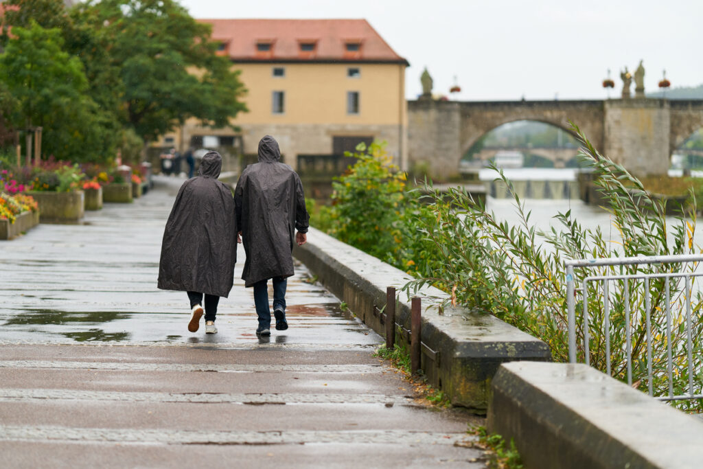 Zwei Menschen mit Ponchos gehen bei Regen an einer Flusspromenade; hinten Steinbrücke und historische Gebäude
