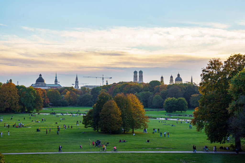 Menschen im Englischen Garten in München mit Blick auf die Skyline und die Türme der Frauenkirche im Hintergrund.