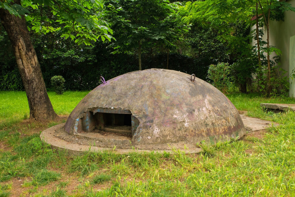 Flacher, halbkugelförmiger Betonbunker mit rechteckiger Öffnung im Lulishte Ismail Qemali Park in Tirana, umgeben von Grasflächen und Bäumen, Tageslicht