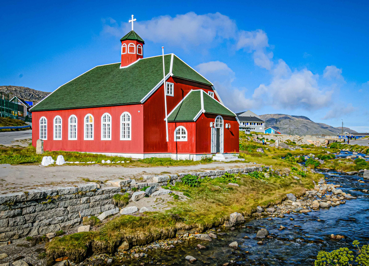 Rote Holzkirche mit grünem Dach an einem Bach; Berge, wenige Häuser, blauer Himmel.