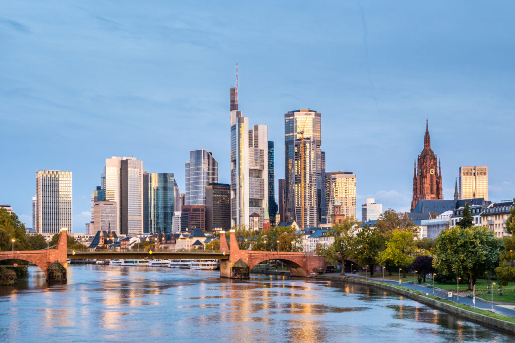Skyline von Frankfurt am Main mit modernen Wolkenkratzern, Brücke und Mainufer bei Sonnenuntergang.