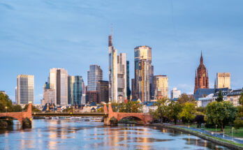Skyline von Frankfurt am Main mit modernen Wolkenkratzern, Brücke und Mainufer bei Sonnenuntergang.