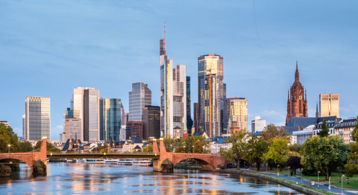 Skyline von Frankfurt am Main mit modernen Wolkenkratzern, Brücke und Mainufer bei Sonnenuntergang.