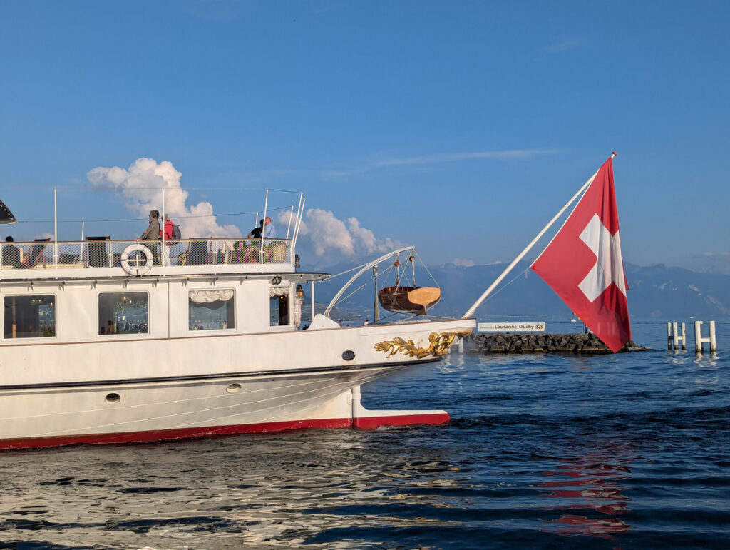Fähre mit Schweizer Flagge nahe dem Anleger Lausanne-Ouchy auf dem Genfersee bei blauem Himmel.