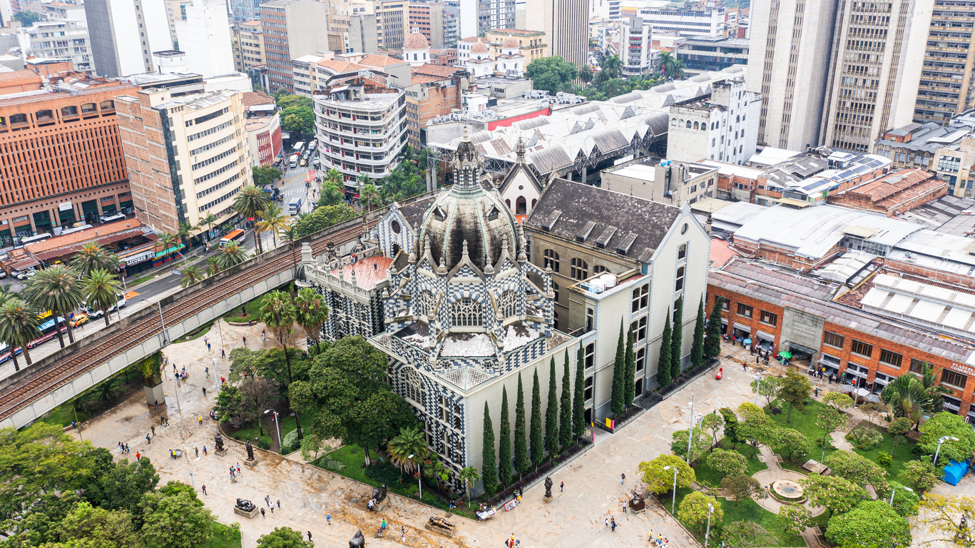 Luftaufnahme der Plaza Botero in Medellín, Kolumbien, mit dem schwarz-weiß gemusterten Palacio de la Cultura Rafael Uribe Uribe, umgeben von Skulpturen, Palmen, Stadtbahntrasse und dicht bebauten Hochhäusern.