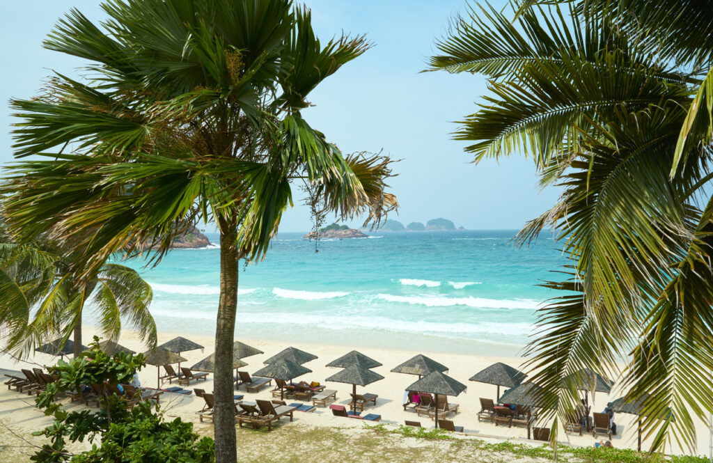 Palmen rahmen einen weißen Strand auf Redang mit Liegestühlen, Sonnenschirmen und türkisfarbenen Wellen
