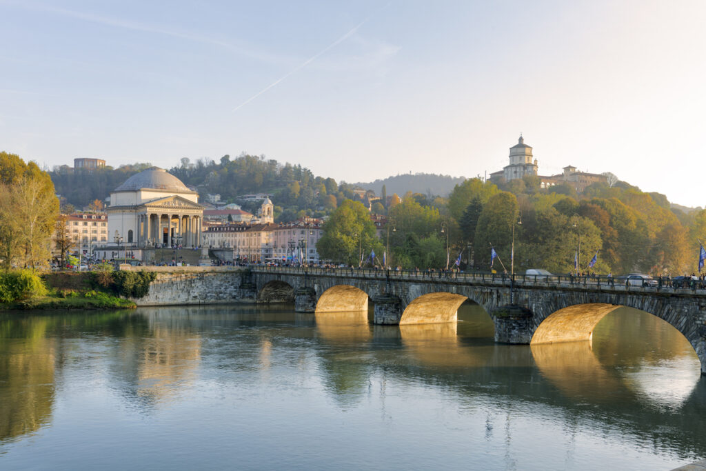 Blick über den Po auf die Ponte Vittorio Emanuele I mit gold schimmernden Bögen, links die Kirche Gran Madre di Dio, rechts am Hügel die Chiesa di Santa Maria del Monte (Monte dei Cappuccini) im weichen Abendlicht.