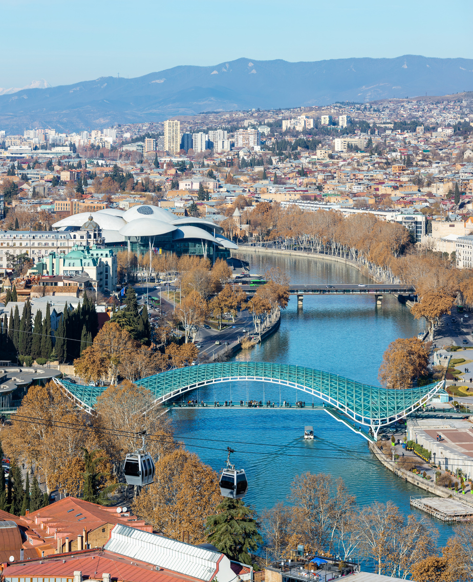 Glasüberdachte Friedensbrücke über die Kura in Tiflis, Seilbahn im Vordergrund, Public Service Hall und Stadtpanorama mit Bergen im Hintergrund.