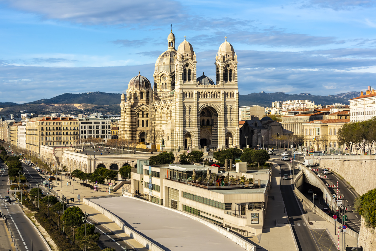 Panorama der Kathedrale La Major in Marseille mit gestreifter Fassade; davor Straßen, moderne Gebäude und Bäume, dahinter Hügel unter blauem Himmel.